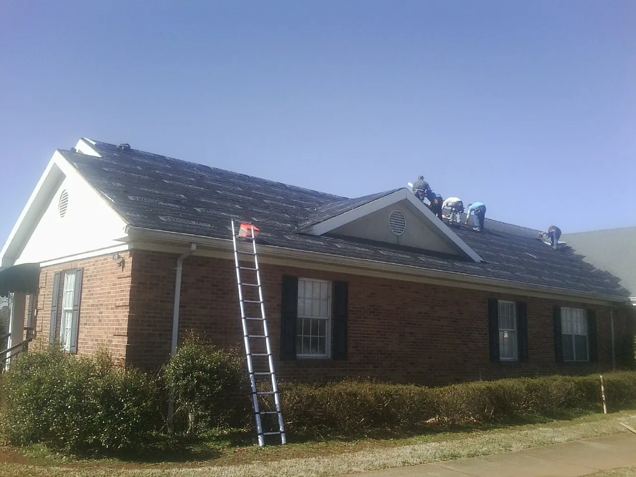 Crew tearing shingles off a brick ranch roof with a ladder set — Metal Roof Repair work by our crew in College Corner