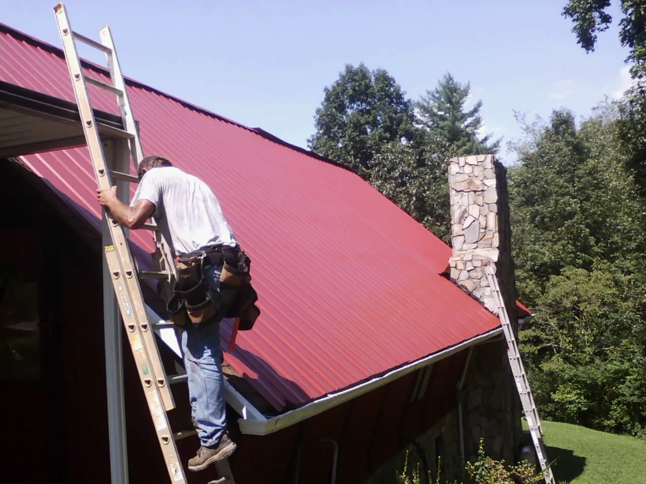 Roofer on a ladder beside a red metal roof and stone chimney — Soffit & Fascia Repair by Roofing Brothers in College Corner