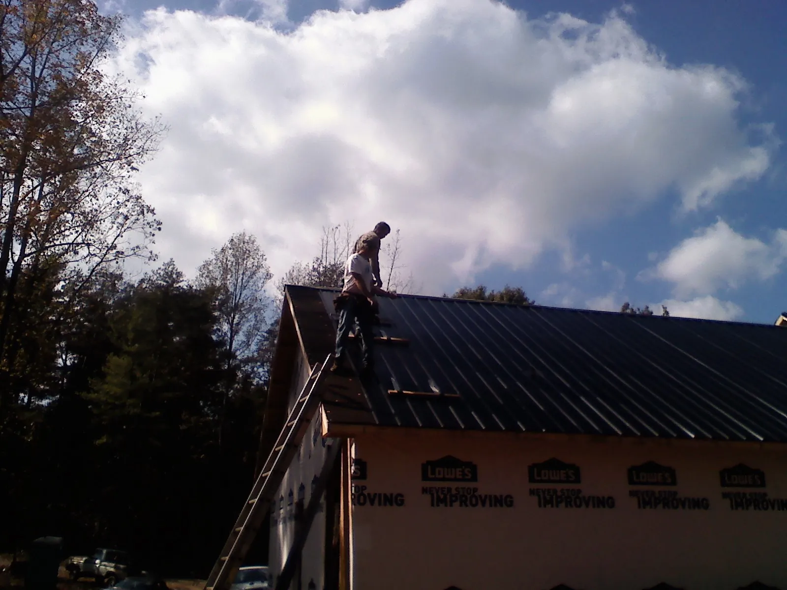 Roofer fastening dark metal panels high against a cloudy sky, from a Roof Inspection job in College Corner